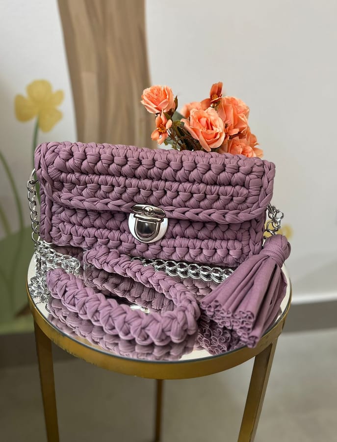 Bolsa de crochê em fio de malha lilás, primeira peça finalizada pela aluna, ao lado de um arranjo de rosas